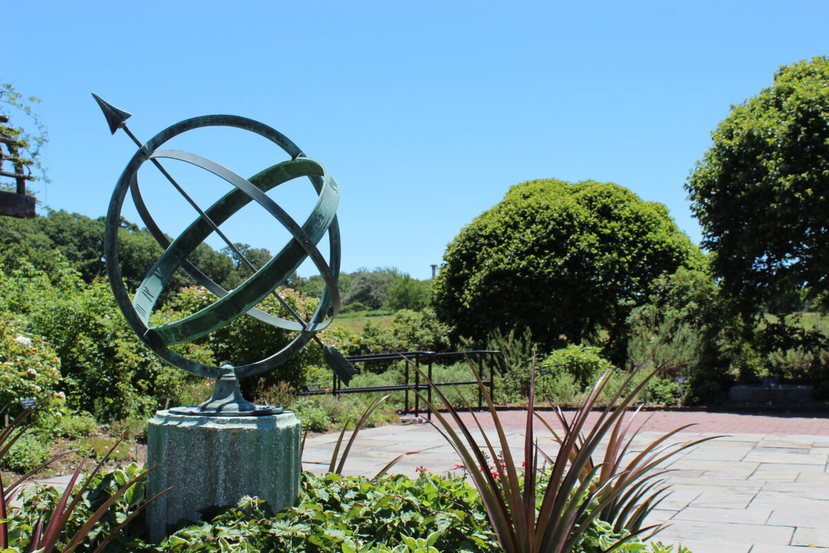 A large green-patinaed copper armilary sphere with an arrow through it is surrounded by plants and trees, as well as a paved walkway.