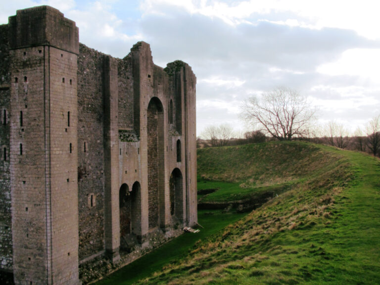 A photo of Castle Rising Castle in England. One wall of the castle takes up the left half of the photo. The right half shows the earthworks ring around the castle. There are trees in the background, bare of leaves because it is winter. The sky is mostly white because it was a pretty cloudy day.