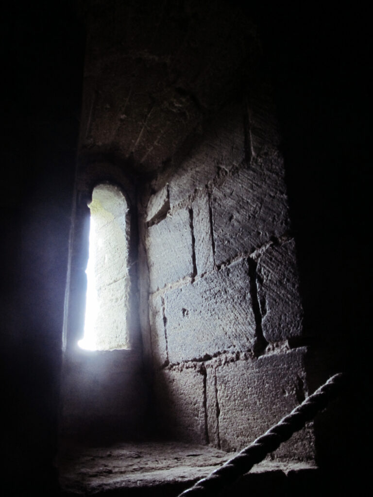 A photo of a narrow window in the thick outer wall of a tower at Castle Rising Castle in England. The light coming through the window is illuminating the niche in the wall where the window is situated. Aside from a bit of thick rope that serves as a railing for a staircase, the rest of the photo is dark.