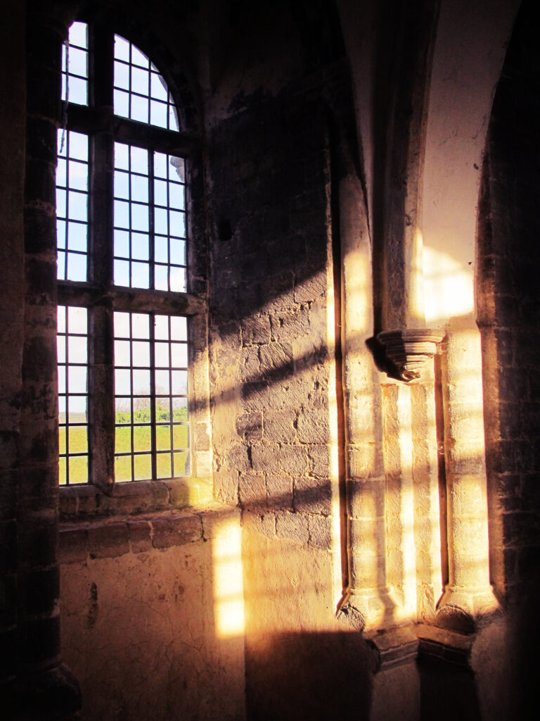 Golden light shines through a large window, illuminating brown and cream stone and plaster in the interior of Castle Rising Castle. Green grass and blue sky can be seen outside the window.