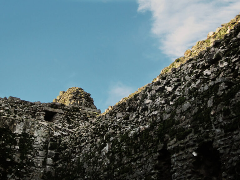 A low-angle shot of the intersection of two partially-ruined walls on the grounds of Castle Rising Castle. There is moss growing on the stones, and rays of sunlight highlight the stones at the top of the walls. The sky is blue, except for a large white cloud in the top right corner.