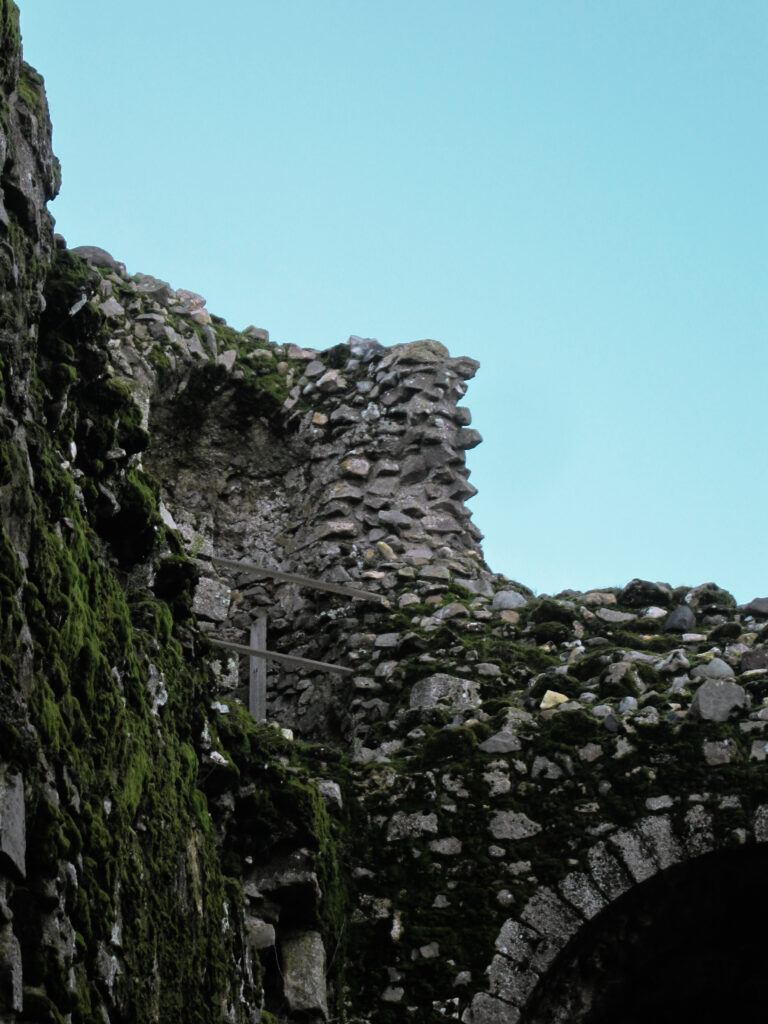 Architectural details such as niches and archways are still discernable in the corner of two partially-ruined stone walls on the grounds of Castle Rising Castle. Moss grows over much of the walls, and portions are supported by wooden beams. The sky in the background is a pale blue.