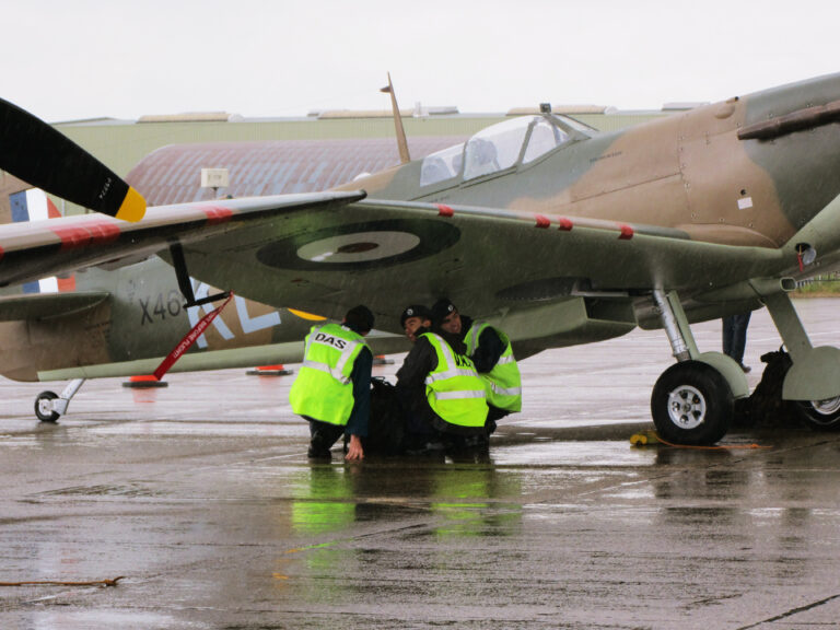 Several men wearing World War II-era RAF uniforms with high visibility vests over them crouch under the wing of a Spitfire plane, sheltering from rain.