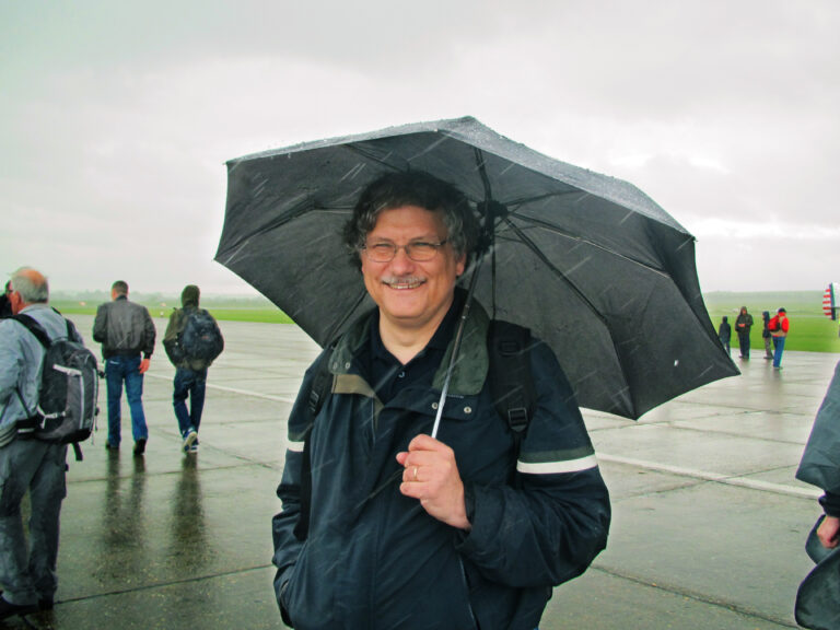 A man stands on a rain-soaked runway, holding an open umbrella. He is wearing glasses and smiling.
