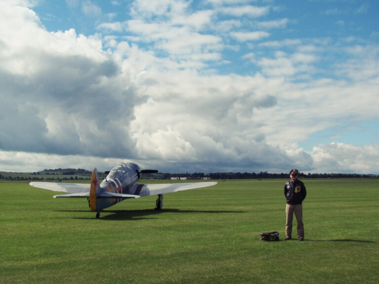 A man in a World War II-era U.S. Army Air Force uniform stands at parade rest in front of the WWII-era single-seat fighter plane he will be flying in an air show later that day.