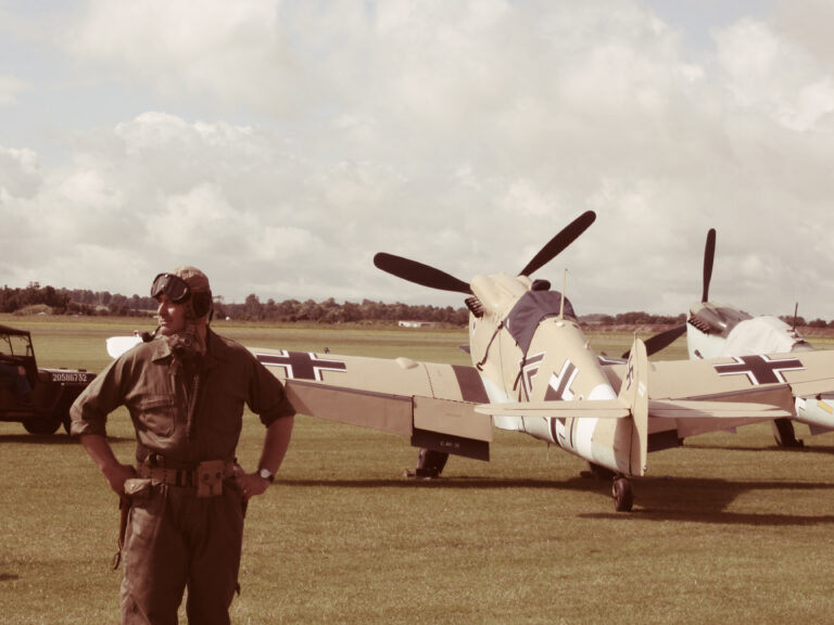 A man in a World War II-era uniform, including a skullcap and flight goggles, smokes a pipe and stands with his hands on his hips. He is standing in front of his WWII-era fighter plane.
