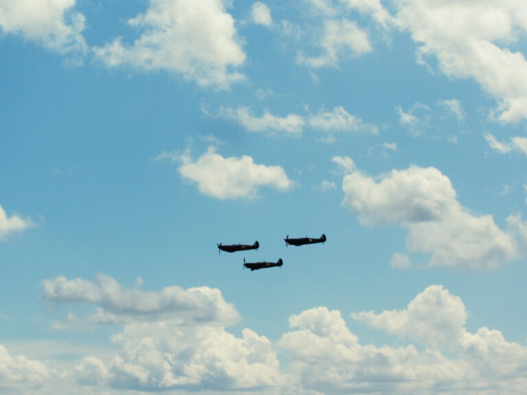 Three WWII-era planes fly in formation in a blue skiy dotted with fluffy white clouds.