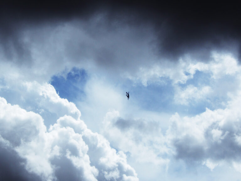 A WWII-era plane is just barely visible in the center of the frame. It is flying past one of the only patches of blue sky visible between fluffy clouds that vary in color from dark grey to white.