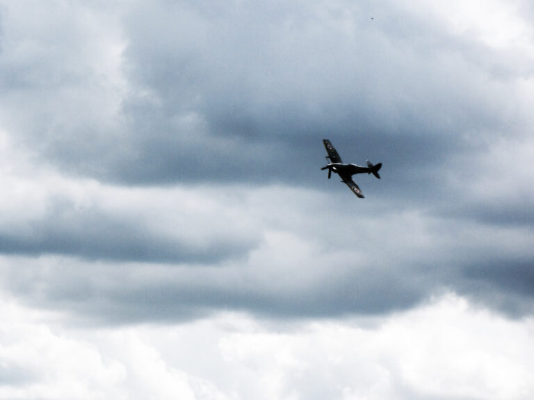 A WWII-era Spitfire fighter plane files past grey and white clouds.
