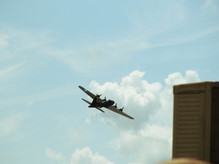 A B-17 bomber banks to the right as it flies past a building in a blue sky dotted with white clouds.