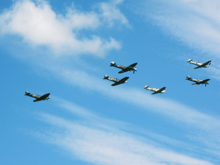 Six WWII-era planes (four Spitfires and two P-51 Mustangs if I am not mistaken) fly in formation through a blue sky with high white clouds.