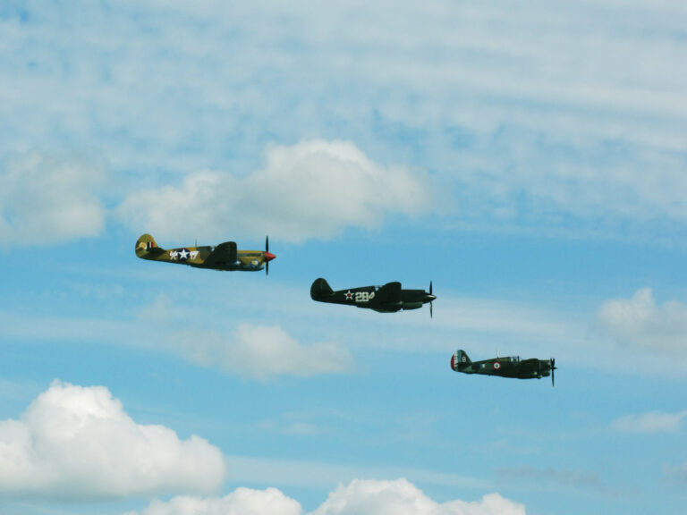 Three WWII-era fighter planes (I believe they are all Spitfires) fly in formation through a blue sky dotted with white clouds.